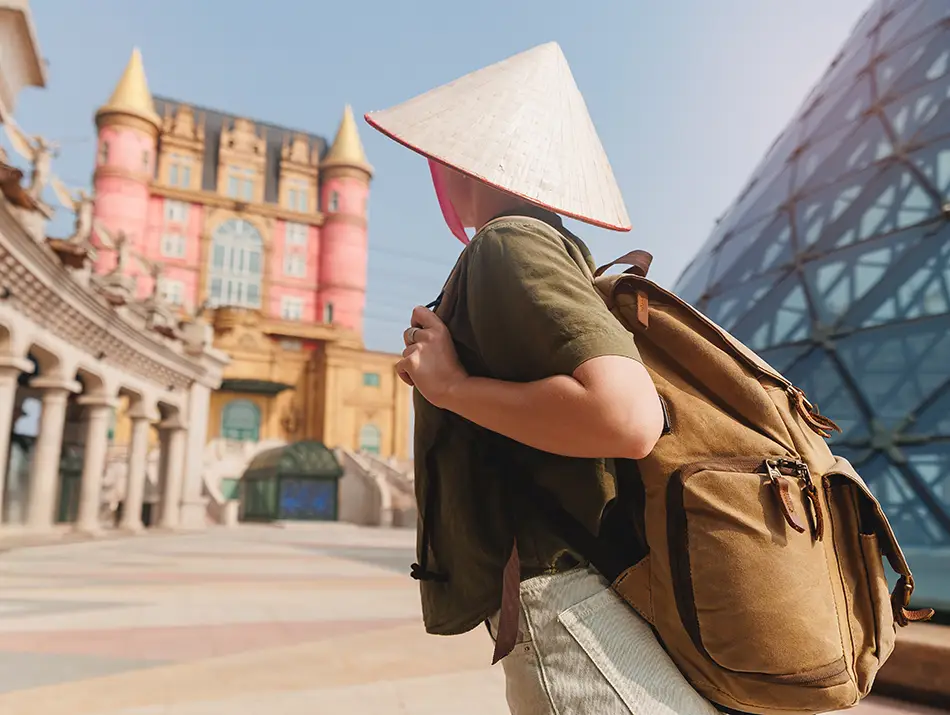 Traveler wearing a conical hat and backpack walks through Ba Na Hills, passing colorful buildings and a glass-domed structure.