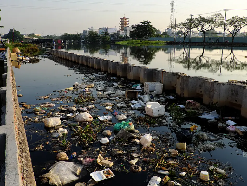 Trash and plastic waste floating in a polluted urban canal, with trees, buildings, and a pagoda visible in the distance.