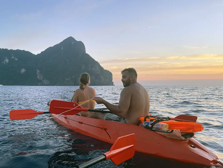 Travelers kayaking on calm water at sunset near a steep island cliff, paddling an orange kayak in open sea.
