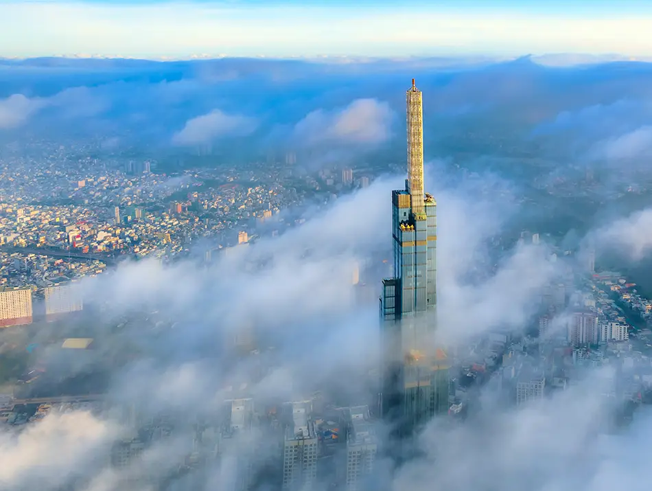 Tall skyscraper rising above low clouds, surrounded by a sprawling cityscape partly covered in mist.