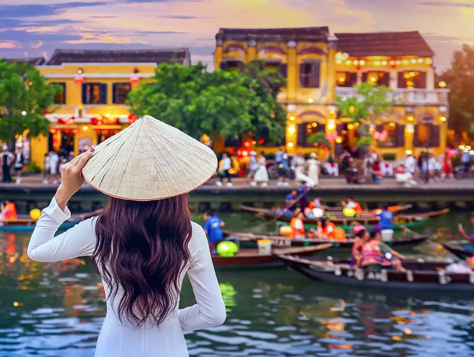 Woman in a white áo dài holds a conical hat while facing Hoi An’s lantern-lit riverside, with boats moving across the water.