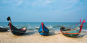 Vietnamese fishing boats painted blue and red rest on a sandy beach with gentle waves and a calm sea in the background.