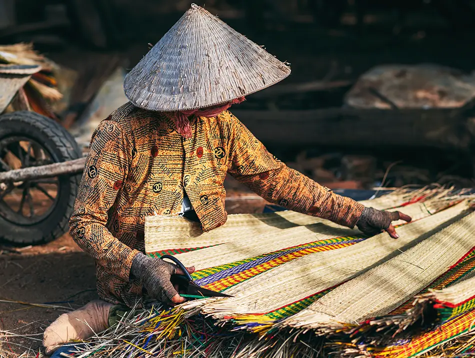 Craftsperson in a conical hat weaves a colorful mat by hand, smoothing reeds on the ground in an outdoor workshop.