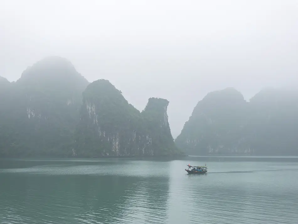 Small fishing boat on calm water surrounded by mist-covered limestone karst islands in Ha Long Bay, Vietnam.