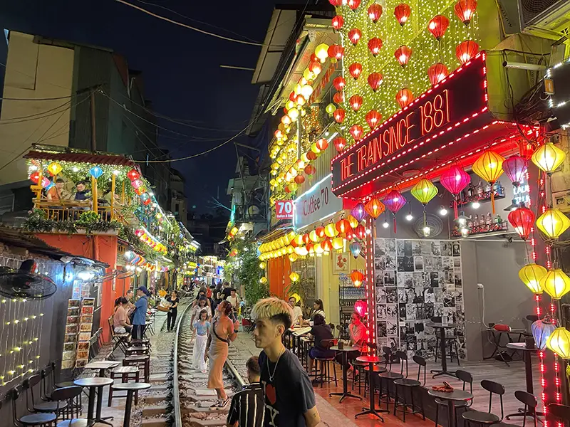 Narrow street in Hanoi with train tracks running between cafes crowded with night visitors.