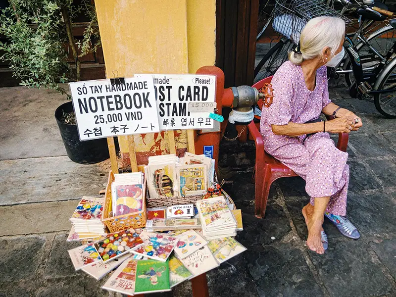 Elderly vendor sitting on the street in Hoi An with handmade notebooks for sale.