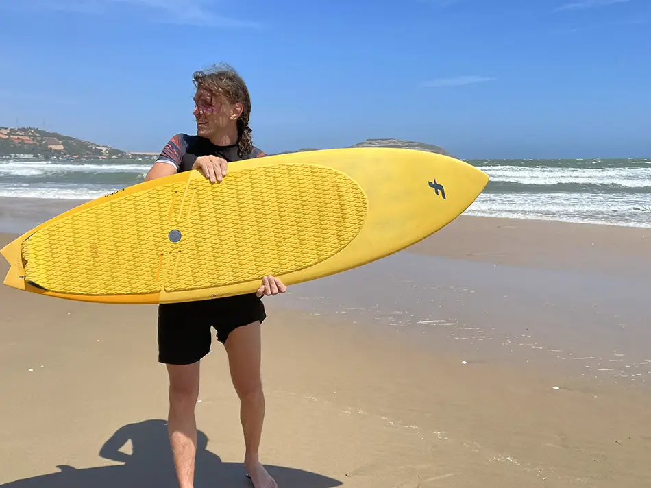 Traveler holding a yellow kite surfboard on a sandy beach in Mui Ne, Vietnam, a popular destination for kitesurfing and coastal travel