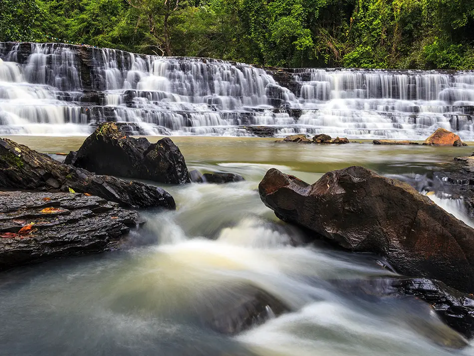 Multi-tiered Thuy Tien Waterfall rushing over rocky ledges, surrounded by green forest in Dak Lak