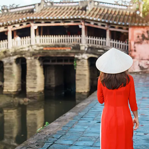 Vietnamese woman in red Ao Dai and conical hat walking toward the Japanese Covered Bridge in Hoi An