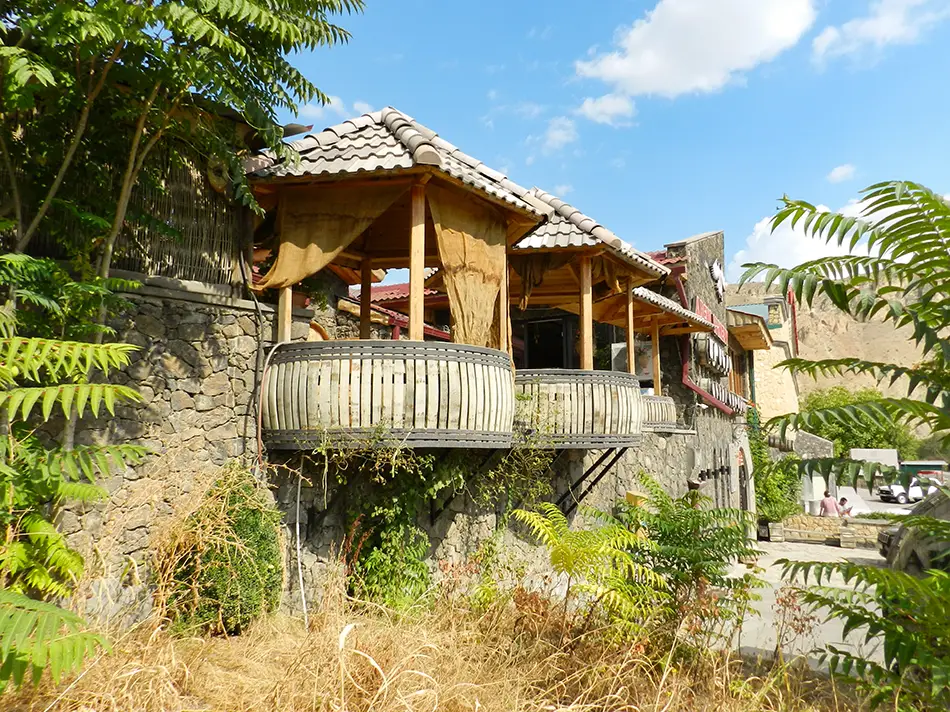 Stone winery with shaded wooden balconies among summer trees on the edge of the village.