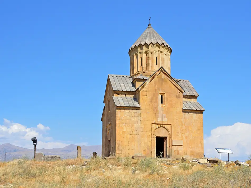Small stone church with conical dome under clear blue sky in a grassy field.