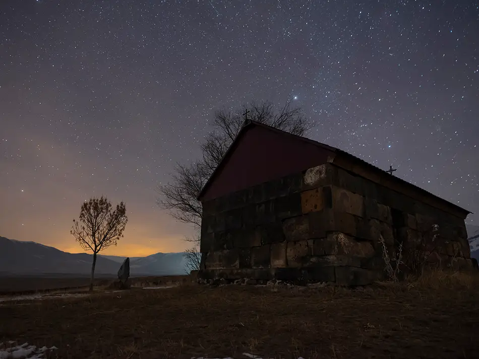 Stone chapel under a star-filled night sky, with mountains faintly visible in the distance.