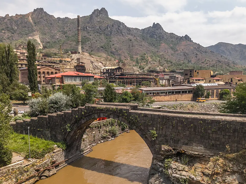 Riverside townscape with bridges, smokestacks, and mountains under hazy afternoon light.