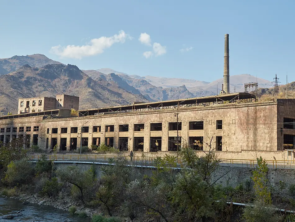 Derelict riverside smelter with tall chimney beneath rugged mountains and blue sky.