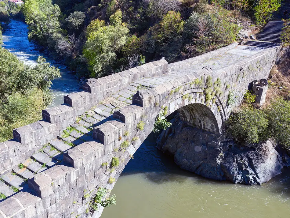 Medieval stone arch bridge spans a forested river gorge with autumn colors.