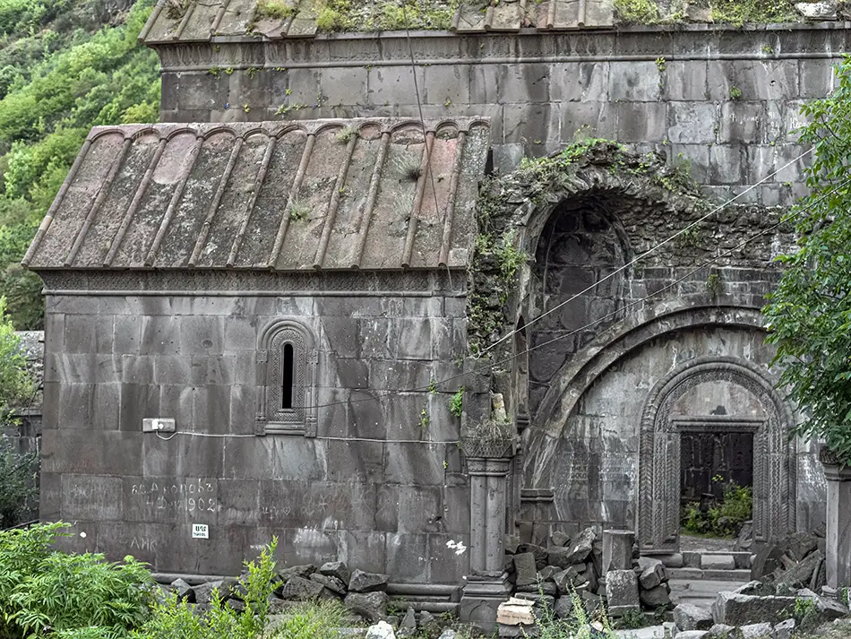 Weathered monastery doorway with intricate carvings set among moss and shrubs.