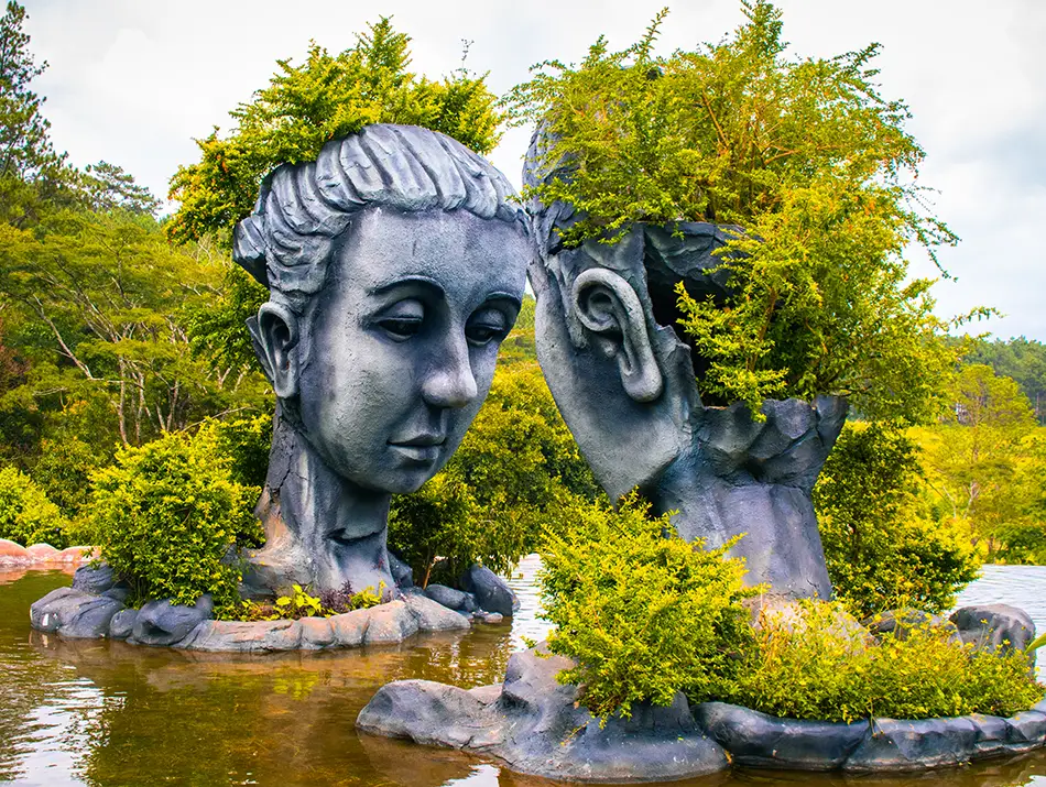 Massive stone-like sculptures of two human heads with trees growing from the tops, reflected in a pond at the Dalat Clay Tunnel Park, one of the most unique attractions in Dalat.