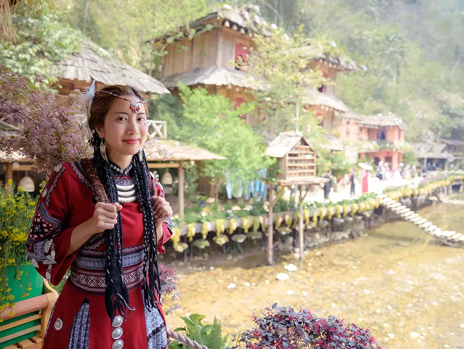 A local woman in traditional clothing stands by wooden riverside homes, reflecting the appeal of visiting Sapa in Vietnam.