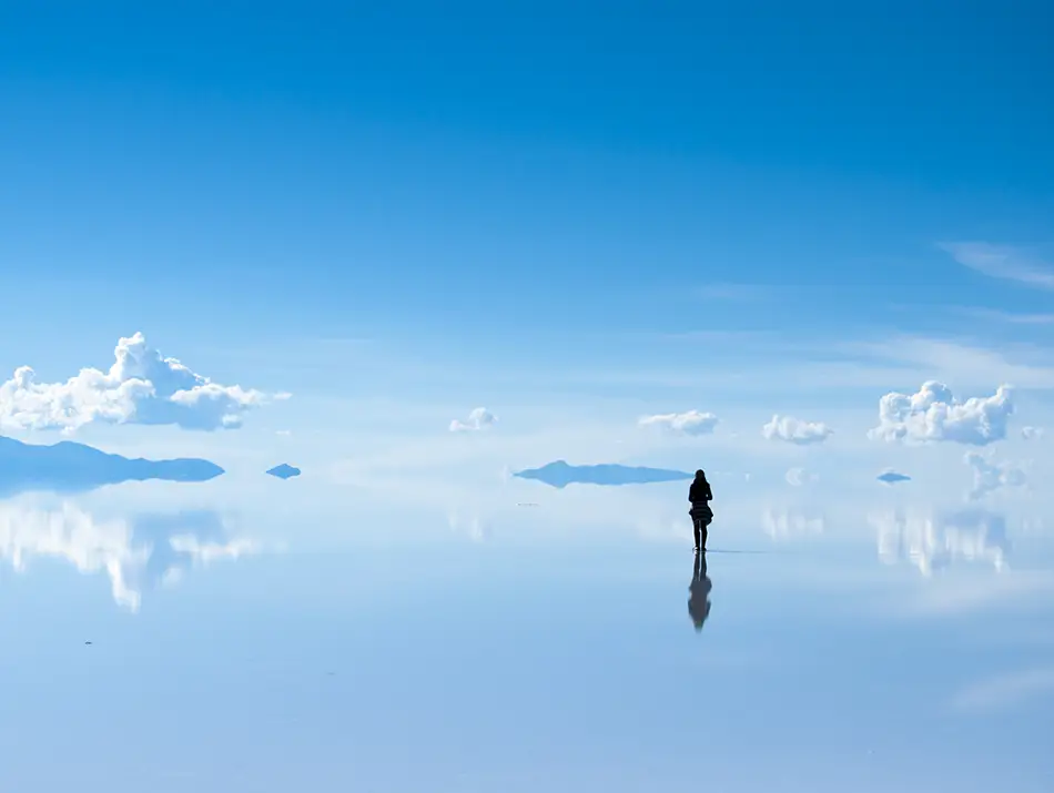 A lone traveler standing on a vast reflective salt flat under clear blue sky.