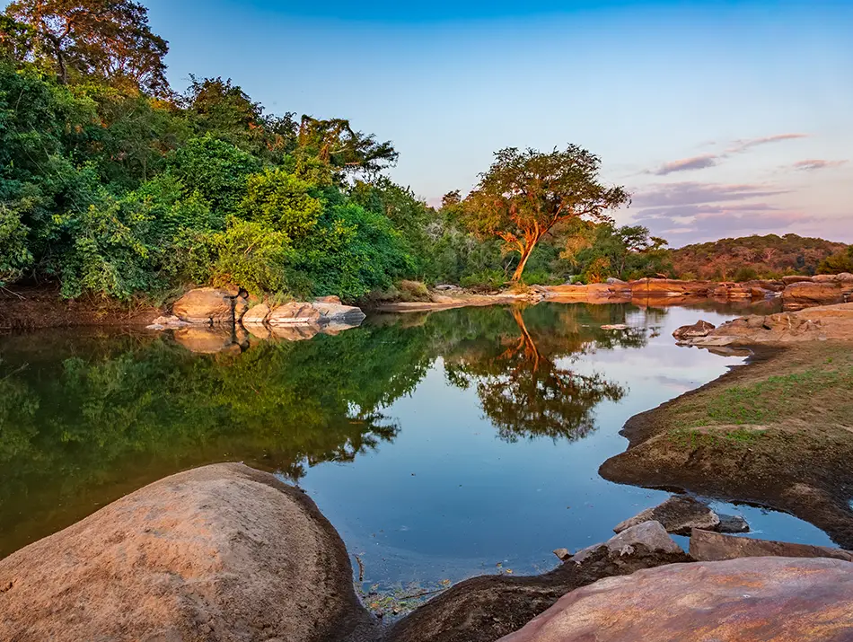 Still river reflecting warm sunset colors with boulders and forest.