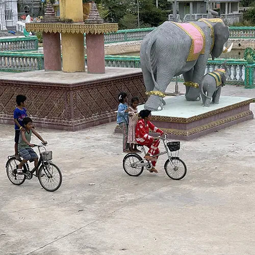 Khmer kids ride past an elephant statue beside the riverside promenade.
