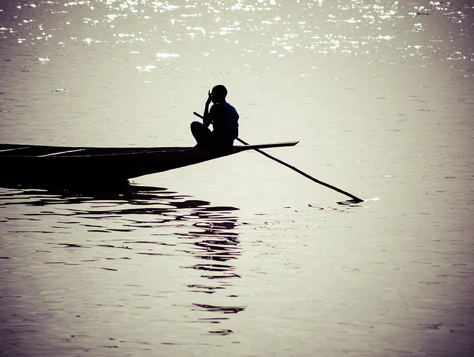 Silhouette of a lone boatman rowing across calm river waters at sunset.