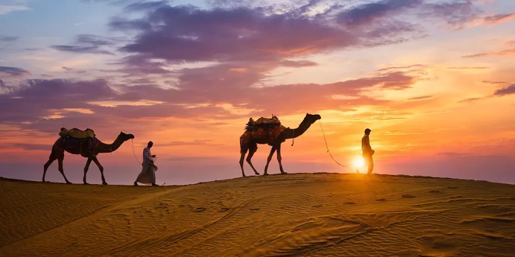 Camel caravan crossing desert dunes at sunset with two travelers walking beside them, illustrating adventure travel in some of the best countries to visit.