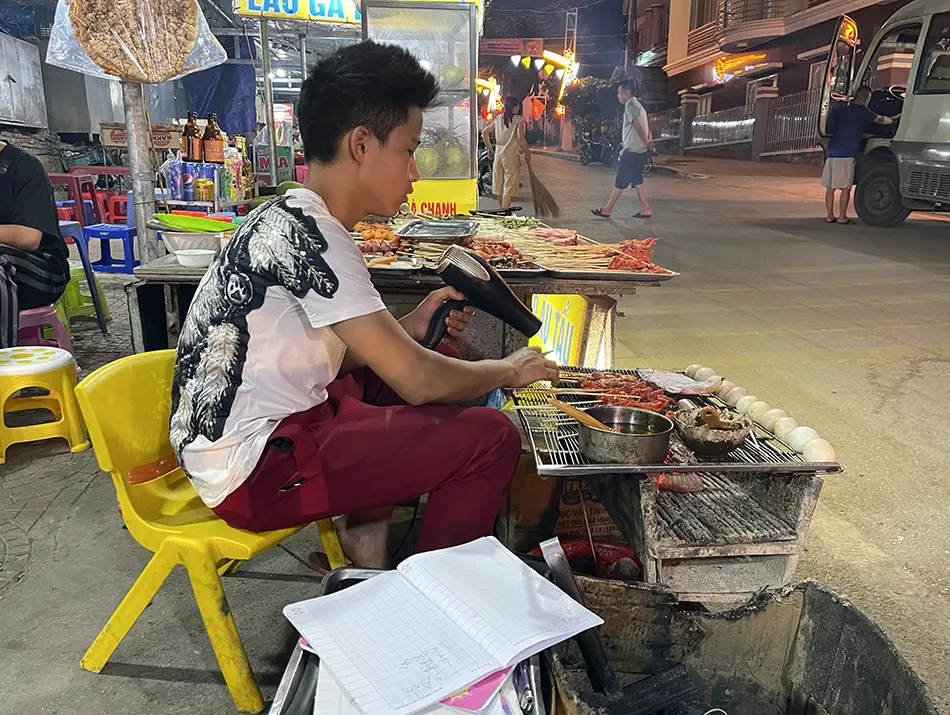 A street vendor blow-drying charcoal grilled skewers demonstrating unique Vietnamese cooking techniques.