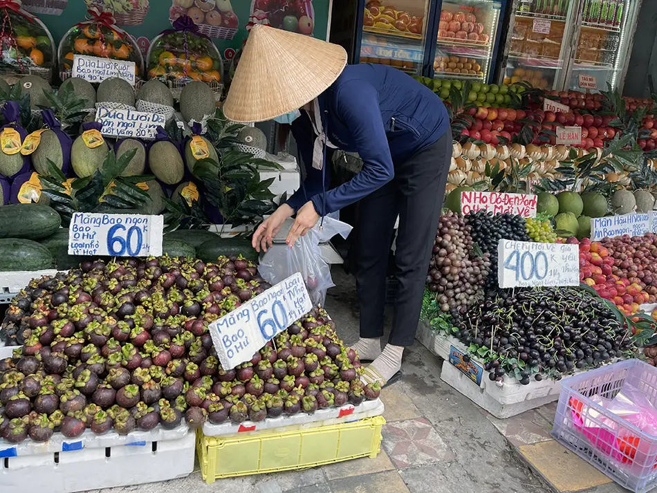Vendor selecting produce at a Vietnamese fruit market, surrounded by mangosteens, grapes, melons, and other fresh seasonal fruits.