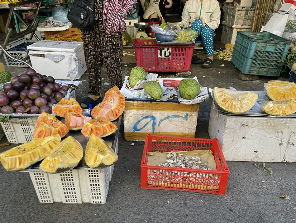 A street market fruit stall displaying sliced jackfruit, whole passion fruits, and several soursop fruits on crates and styrofoam boxes.