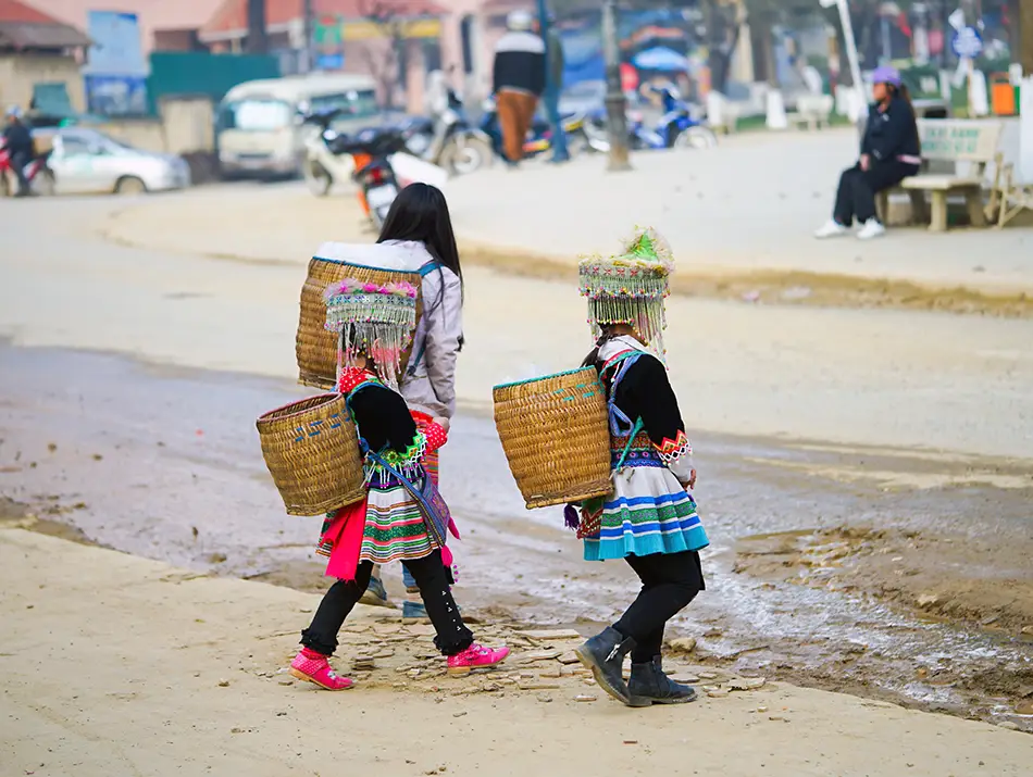 Hmong children carrying woven basket packs walk along a busy roadside near a local market.