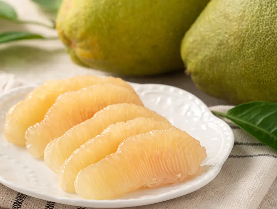 Fresh pomelo segments neatly arranged on a plate, showing their pale, juicy flesh with whole pomelos in the background.