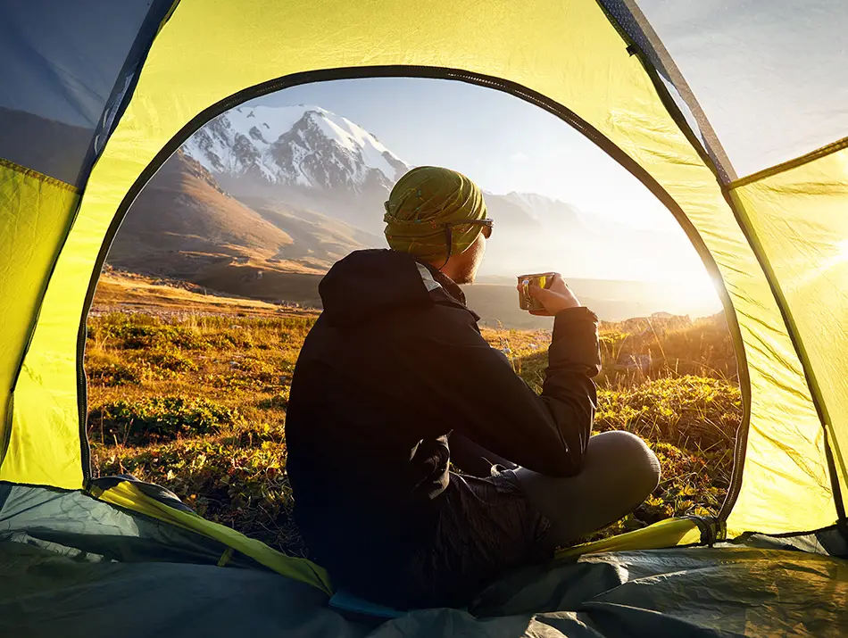 A traveler enjoys morning coffee from a tent overlooking snowy peaks, symbolizing sustainable travel destinations.