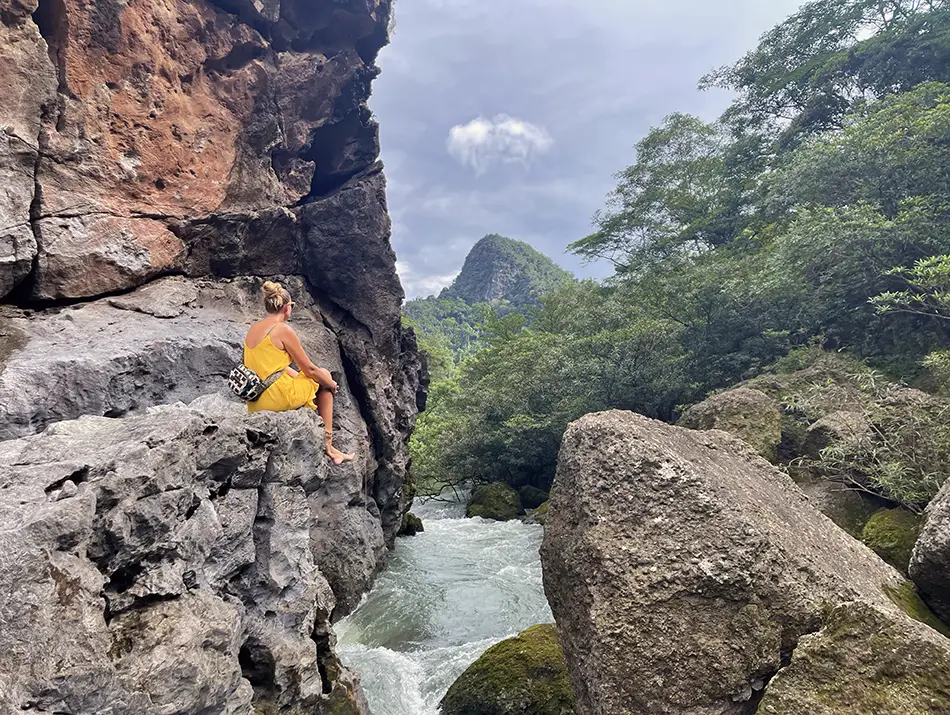 A sustainable traveler sits on a rocky ledge overlooking a fast river and lush mountains.