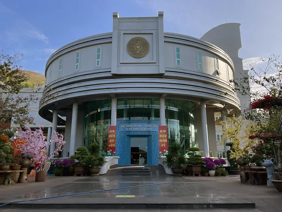 Modern curved museum building with blue glass entrance and decorative flowers out front.