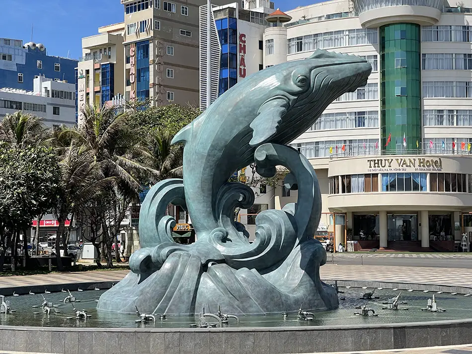 Bronze whale statue leaping above a fountain in a city square with palm trees and buildings behind.