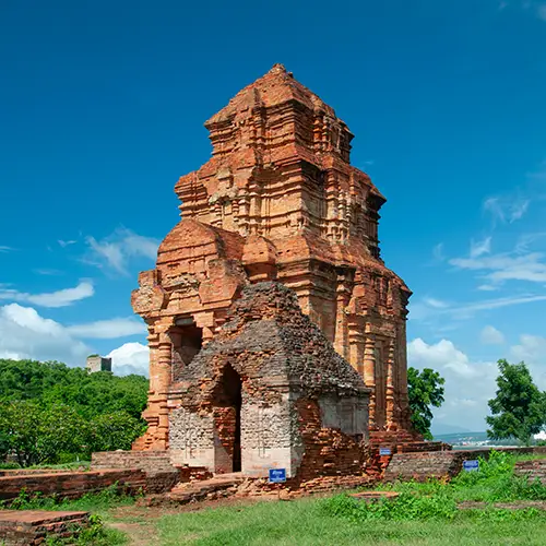 An ancient brick temple tower standing in a grassy clearing beneath a bright blue sky.