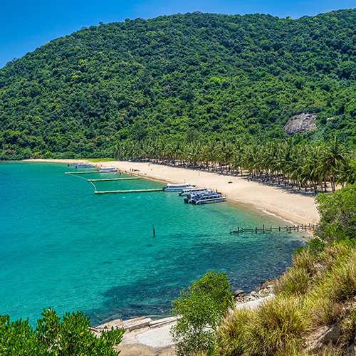 A turquoise bay with boats anchored near a white-sand beach and forested hills, a popular option for a daytrip from Da Nang.