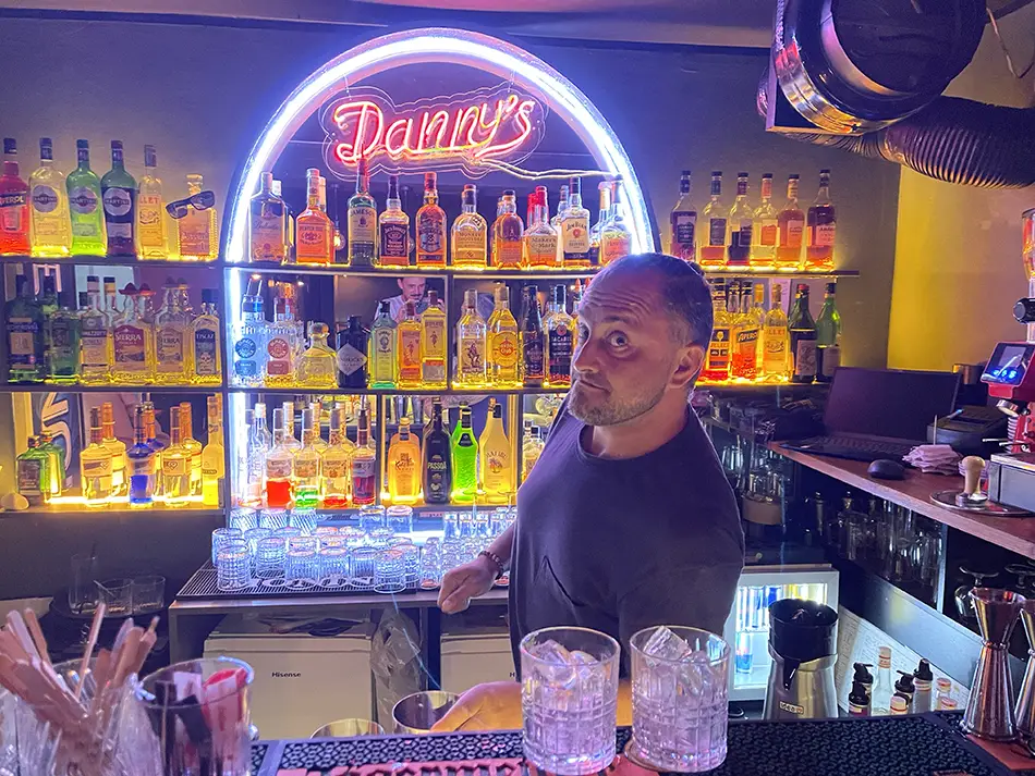 Bartender stands behind a neon-lit bar with shelves of colorful bottles and cocktails.