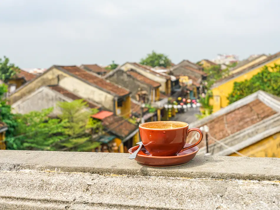 Coffee cup resting on a rooftop ledge overlooking tiled roofs and riverside homes.