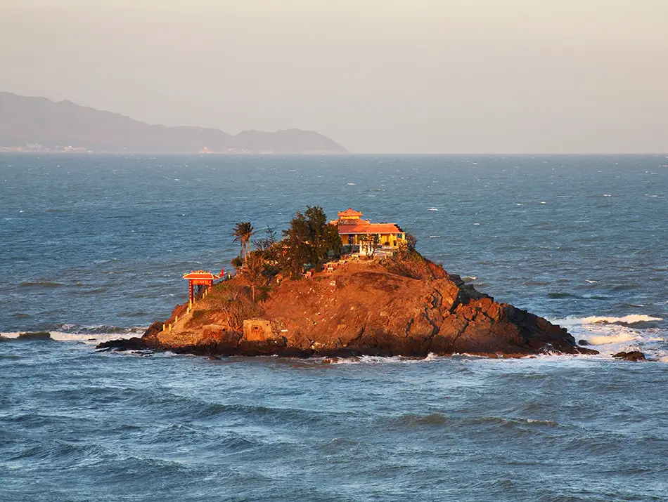 Hon Ba Island and its small hilltop temple surrounded by high tide during sunset, with warm colors reflecting on the water.