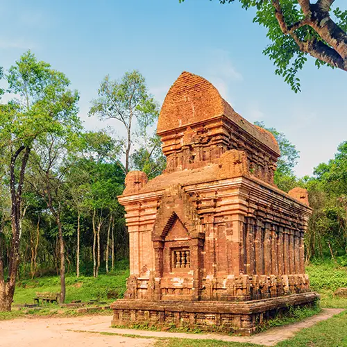 A brick Cham temple tower standing amidst green trees under clear daylight.