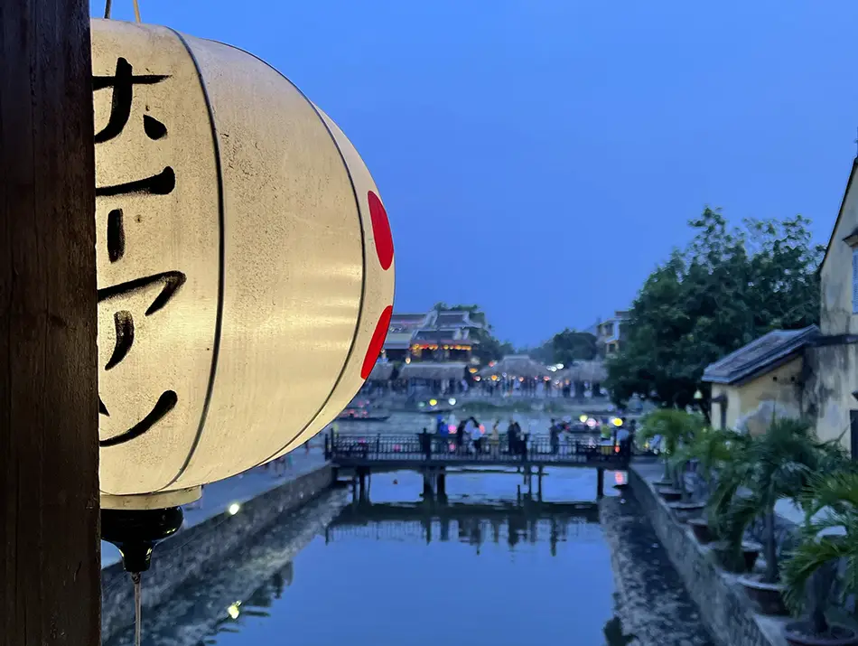 A paper lantern hangs in the foreground overlooking a calm canal and softly lit evening bridge.