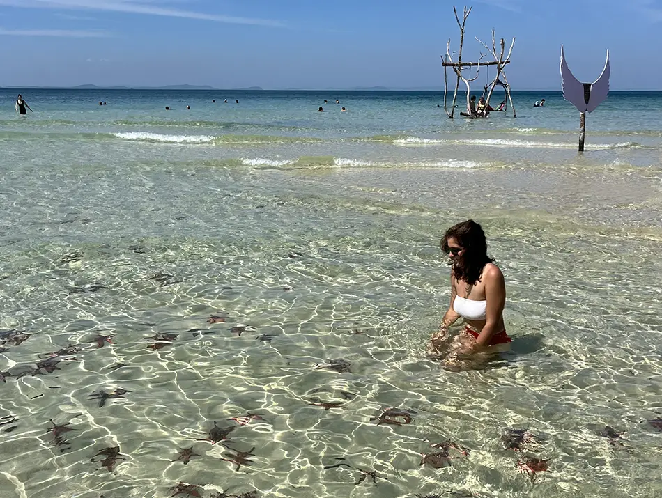 A person stands in shallow, crystal-clear water surrounded by numerous starfish on Starfish Beach.
