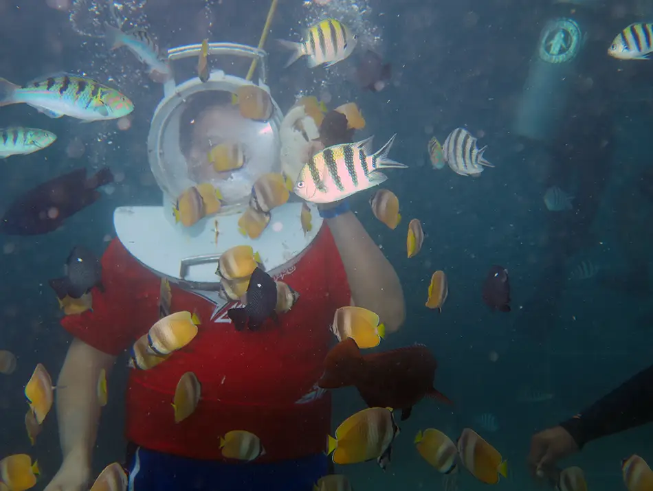 A tourist sea walking on the ocean floor surrounded by tropical fish and coral.