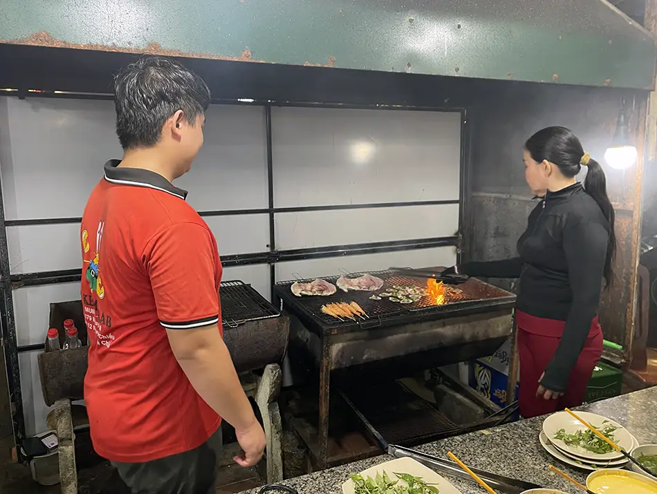 Two people grilling BBQ seafood at a sea-front restaurant, showing the cooking process over an open grill.