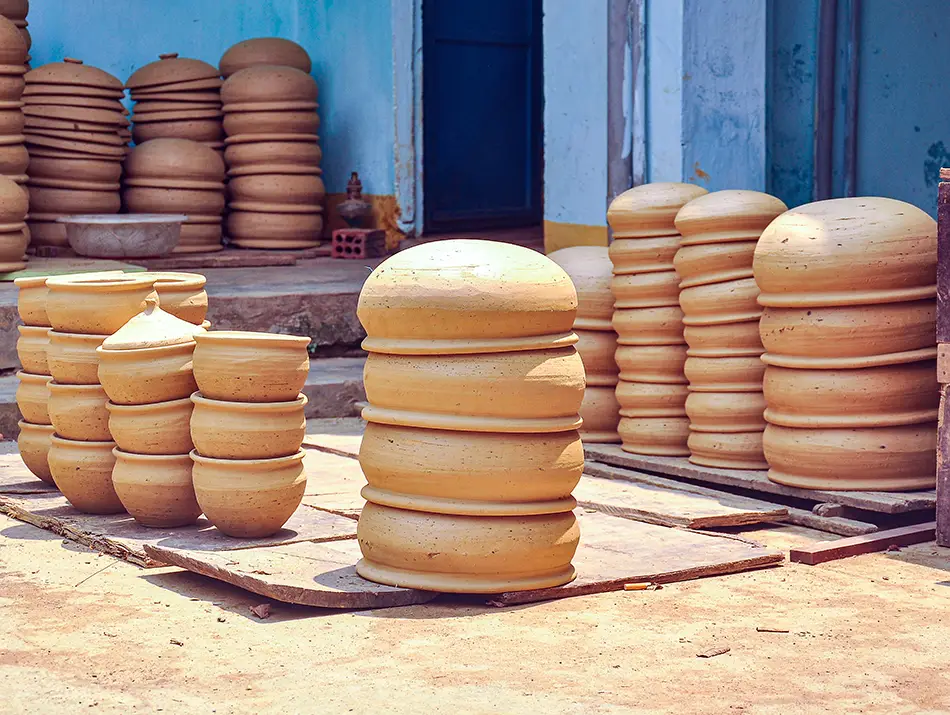 Sun-dried clay pots are stacked in neat rows outside a small workshop, showing traditional pottery preparation in progress.