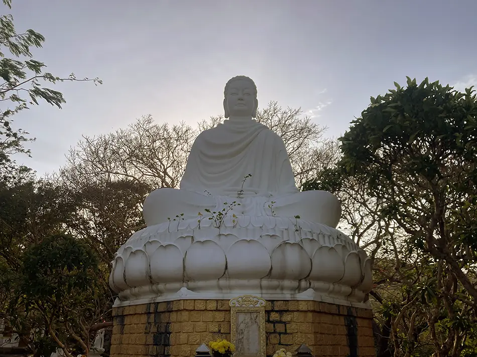 white seated Buddha statue framed by trees against a bright sky.
