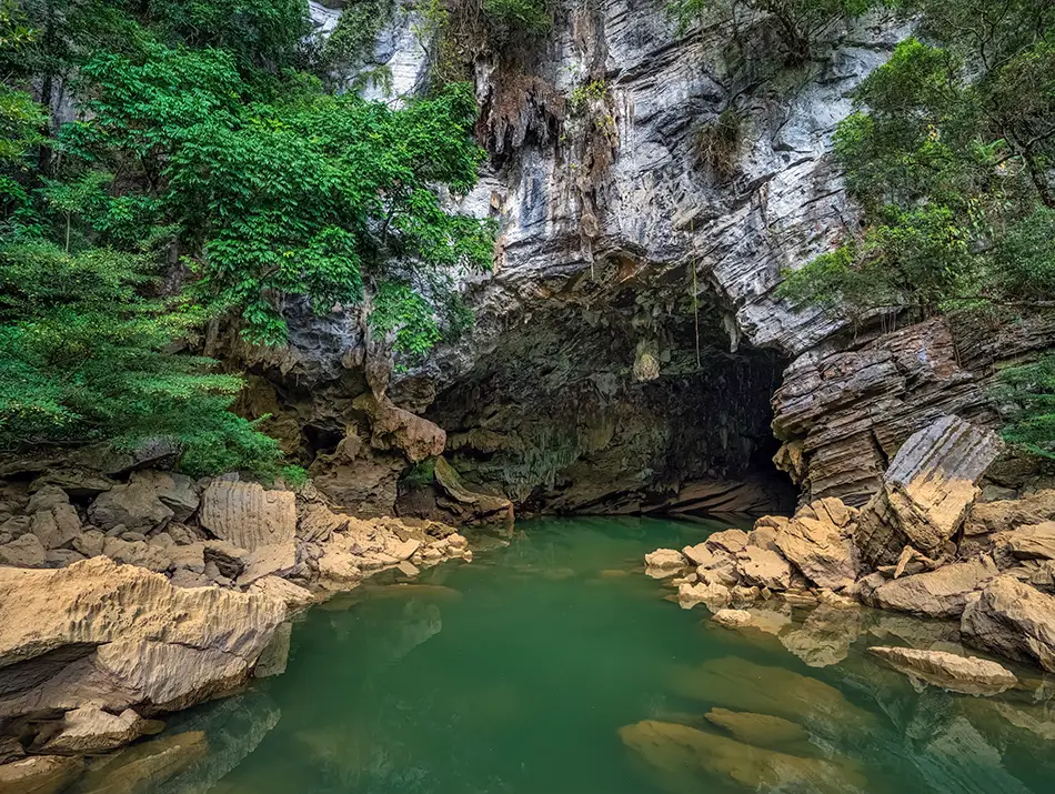 Rocky river entrance surrounded by forest, leading into a remote limestone cave system.