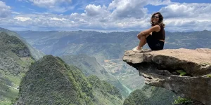 Traveler sits on a rocky ledge above dramatic green mountains and clouds.